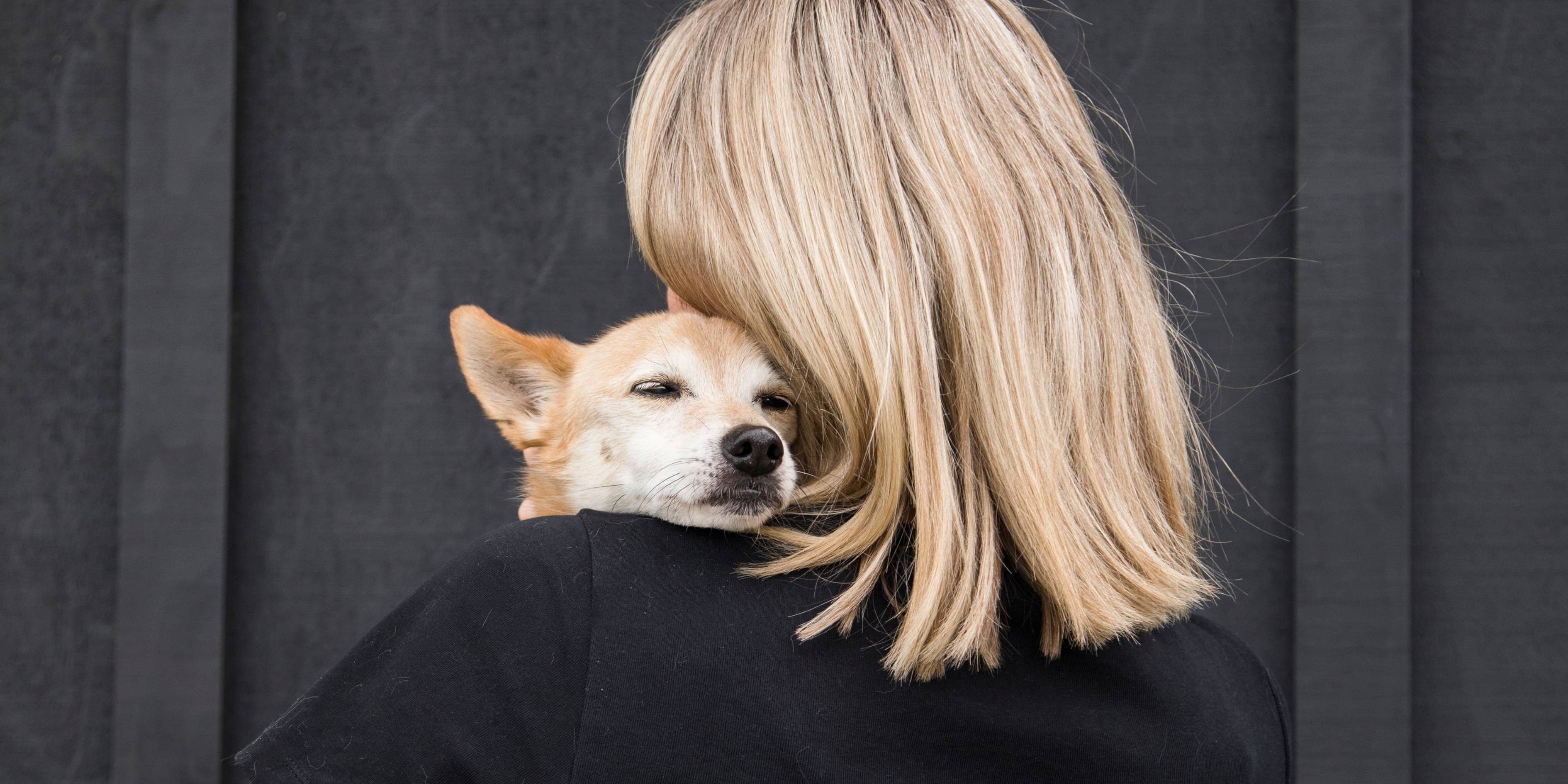A blonde woman holds a small, tan dog with closed eyes on her shoulder.